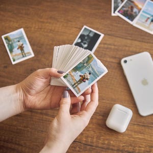 K&ouml;nnte beinhalten: Ein Stapel kleiner, rechteckiger Fotoabz&uuml;ge mit Outdoor-Szenen, gehalten in H&auml;nden mit manik&uuml;rten N&auml;geln. Ein wei&szlig;es Smartphone und ein kabelloses Kopfh&ouml;rer-Etui liegen auf einer Holzoberfl&auml;che, zusammen mit anderen verstreuten Fotos.