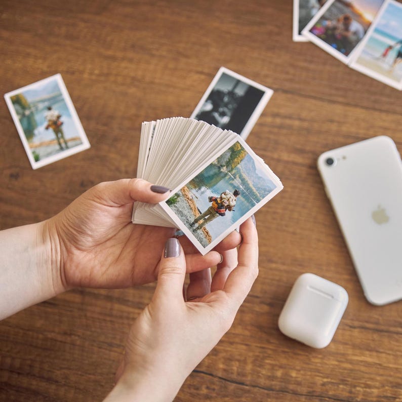 K&ouml;nnte beinhalten: Ein Stapel kleiner, rechteckiger Fotoabz&uuml;ge mit Outdoor-Szenen, gehalten in H&auml;nden mit manik&uuml;rten N&auml;geln. Ein wei&szlig;es Smartphone und ein kabelloses Kopfh&ouml;rer-Etui liegen auf einer Holzoberfl&auml;che, zusammen mit anderen verstreuten Fotos.