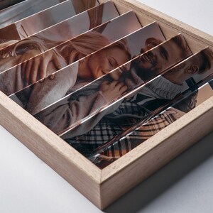 May include: Wooden photo display with multiple angled slots, each holding a photograph. The photos depict a couple embracing and smiling. The frame is made of light-colored wood, and the photos are visible through the angled slots.