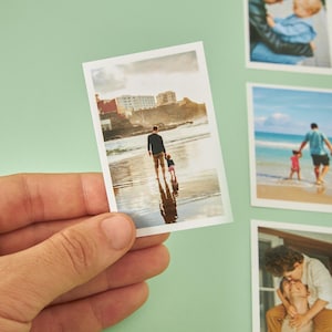 K&ouml;nnte beinhalten: Ein kleines, rechteckiges Foto zeigt einen Mann und ein Kind, die am Strand spazieren gehen. Der Mann tr&auml;gt ein dunkles Hemd und khakifarbene Hosen, das Kind ist in Rosa gekleidet. Das Foto wird in einer Hand vor hellgr&uuml;nem Hintergrund gehalten.