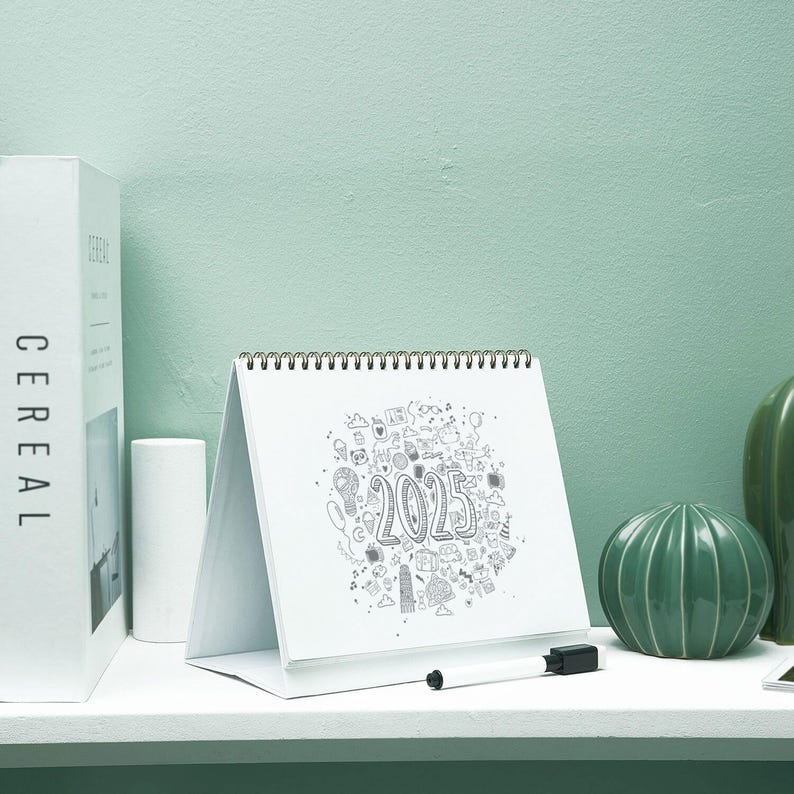 May include: A white desk calendar for the year 2025, featuring a spiral binding and a stand. The calendar has a doodle design around the numbers. A black marker rests on the stand. A white box labeled "CEREAL" is on the left.