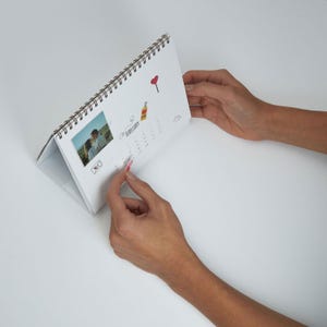 May include: A white desk calendar with a spiral binding, featuring a photo and illustrations. The calendar is open to February, with a small heart sticker being placed on a date. The calendar has a photo of a couple.