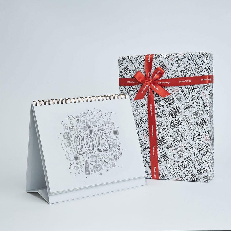 May include: A white desk calendar for 2025 with a spiral binding, featuring a doodle design. Beside it is a gift box wrapped in patterned paper and tied with a red ribbon. The gift box has a red ribbon with the word "Developer" printed on it.