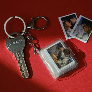 May include: A silver key with the word "KALE" engraved on it, attached to a keychain with a small photo album. The album contains several small photos. Two additional photos are next to the album on a red surface.