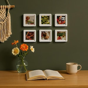 May include: Six white square photo frames with different images are mounted on a dark green wall. A vase of orange and yellow flowers, an open book, and a beige mug sit on a wooden table. A macrame wall hanging is on the left.