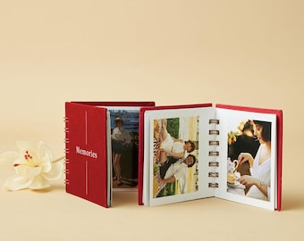 May include: Red photo album with the word "Memories" printed on the cover. The album is open, displaying several photographs. The album is spiral-bound and has a white flower in the foreground.