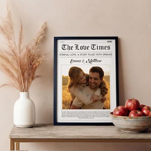 May include: A framed print titled "The Love Times" with a photo of a couple embracing. The print has a black frame and is displayed on a wooden table. A white vase with dried flowers and a bowl of red apples are also on the table.