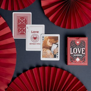 May include: A deck of playing cards with a red and white design, featuring the word "LOVE" and the phrase "always wins." Several individual cards are displayed, including one with a photo of a couple kissing. Red paper fans are arranged around the cards.