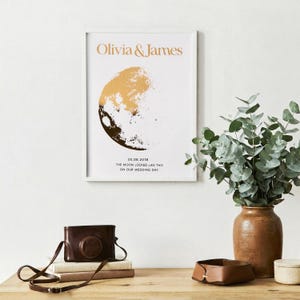 Pode incluir: Uma impressão emoldurada com os nomes "Olivia & James" em letras douradas. A obra de arte retrata uma fase lunar dourada e preta. Abaixo, o texto diz "05.08.2018 THE MOON LOCKED LIKE THIS ON OUR WEDDING DAY."