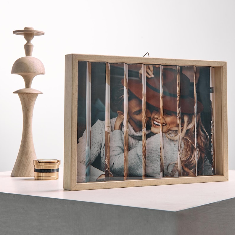 May include: A wooden photo frame with a picture of a couple, cut into vertical strips. The frame is light-colored, and the photo shows a man and woman smiling. A wooden decorative object and a gold and black container are also visible.