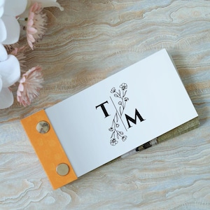 Può includere: Un biglietto bianco con un disegno floreale nero e le lettere "T" e "M" in un monogramma diviso. Il biglietto è rilegato con un cinturino in pelle arancione brillante e due rivetti in ottone.