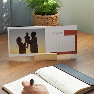 May include: A desktop calendar for August, featuring a family silhouette photo. The calendar is supported by a wooden block. A person is writing in a notebook with a pen. A small potted plant sits in the background.