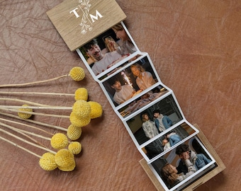 May include: A wooden accordion photo album with a light brown finish, featuring the letters T and M with a floral design on the cover. The album unfolds to display a series of candid photographs of people, accompanied by a cluster of dried yellow flowers.