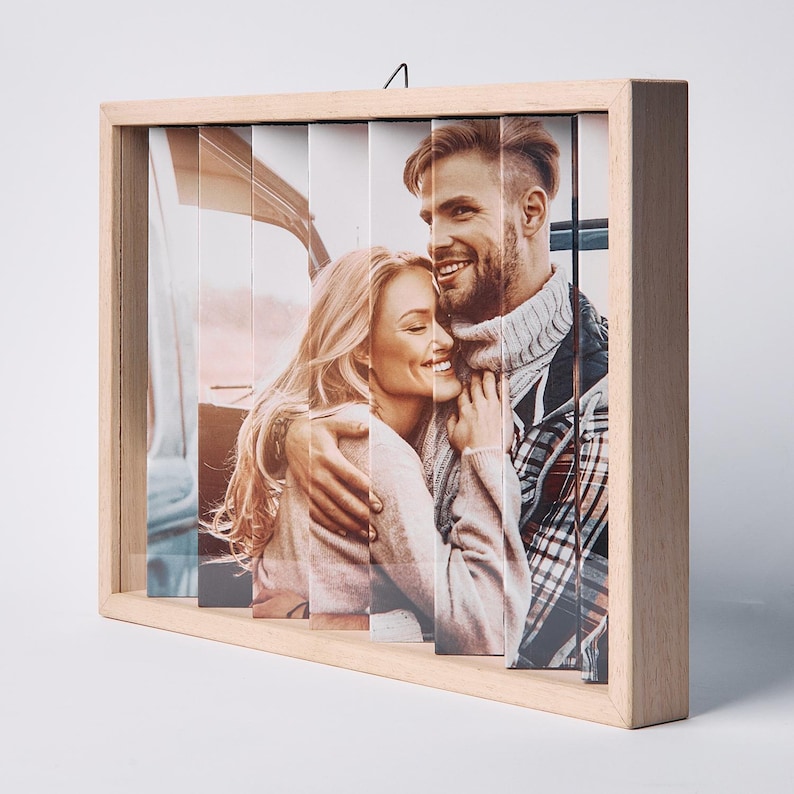 May include: A wooden photo frame with a unique design. The frame holds a photo split into vertical strips, creating a layered effect. The image features a smiling couple embracing. The frame is made of light-colored wood.