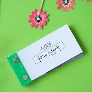 May include: A white booklet with a green spine and silver metal fasteners. The front cover features a simple floral design and the text "Jane & Jack, 05.07.2016".