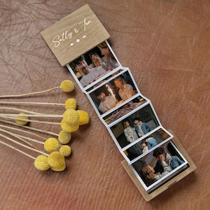 Könnte beinhalten: Eine Fotobox aus hellem Holz mit einem ausziehbaren Akkordeonstreifen aus sechs Fotos. Der Holzdeckel ist mit dem Text Sally & Tom graviert. Ein Bündel getrockneter gelber Trommelstöckchen-Blüten liegt neben dem Fotodisplay.