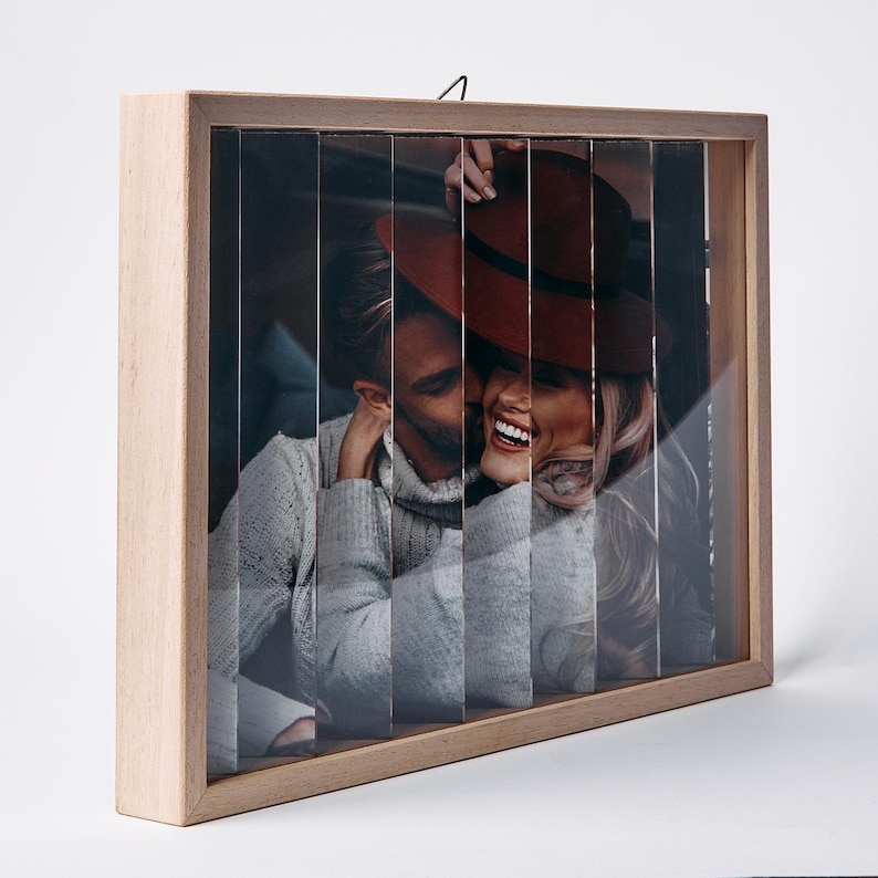 May include: A wooden framed photo with a unique design. The image is split into vertical strips, creating a layered effect. The photo features a couple, with one wearing a red hat. The frame is made of light-colored wood.
