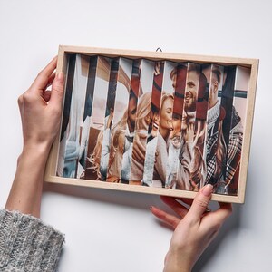 May include: A wooden framed photo with a unique, layered design. The image is divided into vertical strips, creating a 3D effect. The photo features a group of people, with a focus on their faces and expressions. The frame is held by hands.