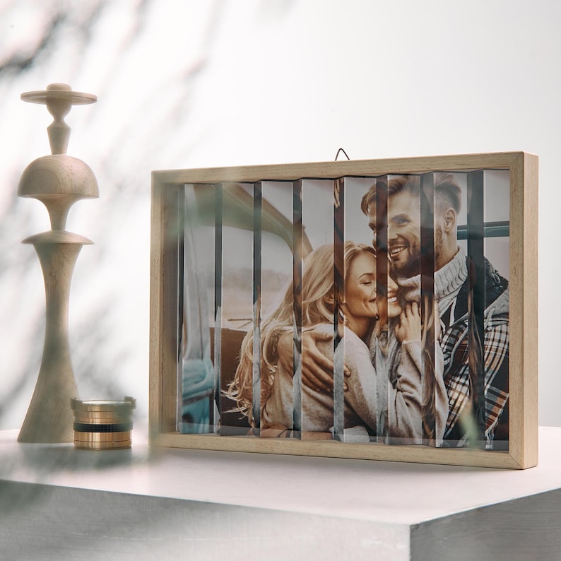May include: A wooden photo frame with a unique design. The frame holds a photo of a couple, divided into vertical strips. A decorative wooden candlestick and a small gold and black ring holder are on the left.