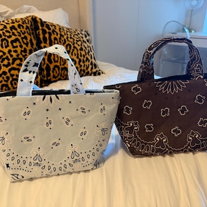 May include: Two small tote bags with handles, one in light blue and one in dark brown. Both bags feature a white paisley bandana print pattern with black accents. The bags have a structured, open-top design and are made of fabric.