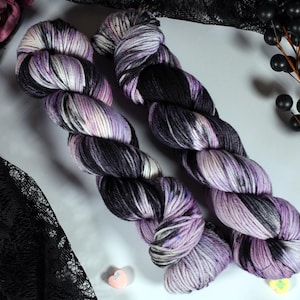 May include: Two skeins of hand-dyed yarn in shades of purple, black, and white. The yarn is arranged on a white surface with decorative elements including purple roses, black lace, and black berries.