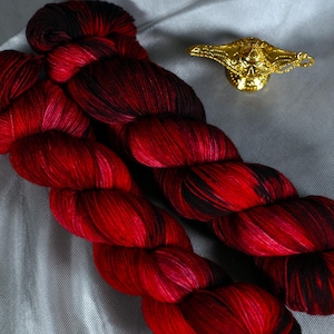 May include: Two skeins of hand-dyed yarn in shades of red and black, with a metallic gold genie lamp-shaped charm. The yarn is arranged on a gray fabric, showcasing the vibrant color variations.