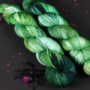 May include: Two skeins of green and white yarn with a speckled pattern. The yarn is arranged in a criss-cross pattern.