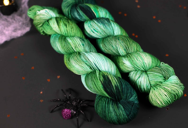 May include: Two skeins of green and white yarn with a speckled pattern. The yarn is arranged in a criss-cross pattern.