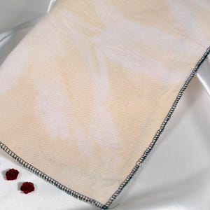 Può includere: Un tessuto beige con un bordo cucito nero è esposto su una superficie di raso bianco. Il tessuto presenta un motivo sottile ed è accompagnato da tre piccole decorazioni di rose rosse.