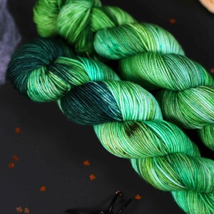 May include: Two skeins of green and black yarn with a speckled pattern. The yarn is arranged in a loose coil.