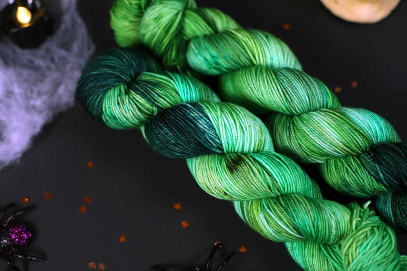 May include: Two skeins of green and black yarn with a speckled pattern. The yarn is arranged in a loose coil.