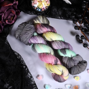May include: A colourful skein of hand-dyed yarn in a gradient of colours, including grey, pink, yellow, and mint green. The yarn is arranged on a white surface with purple flowers and black berries in the background. Ideal for knitting or crochet projects.