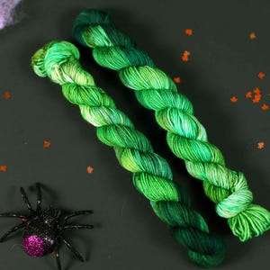May include: Two skeins of green yarn with a gradient of colour from light green to dark green. The yarn is twisted together and is lying on a dark green surface.