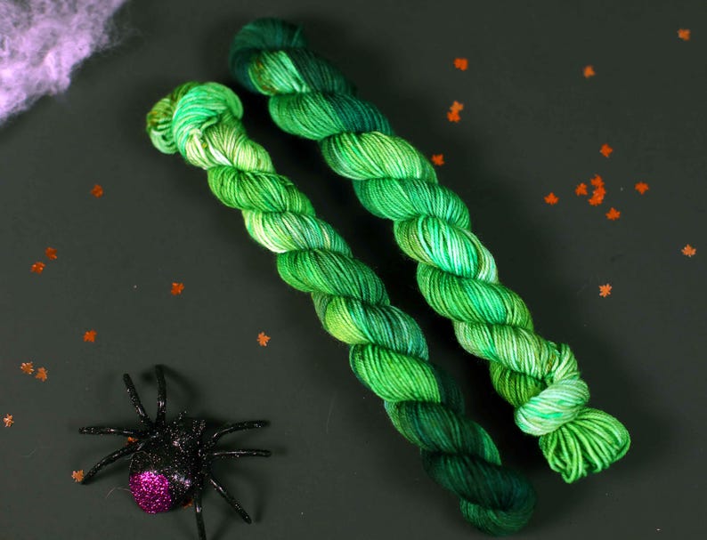 May include: Two skeins of green yarn with a gradient of colour from light green to dark green. The yarn is twisted together and is lying on a dark green surface.