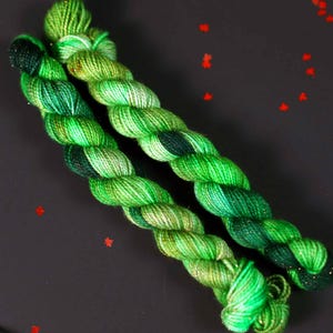 May include: Two skeins of green yarn with a speckled pattern. The yarn is twisted together and has a dark green and light green colour variation.