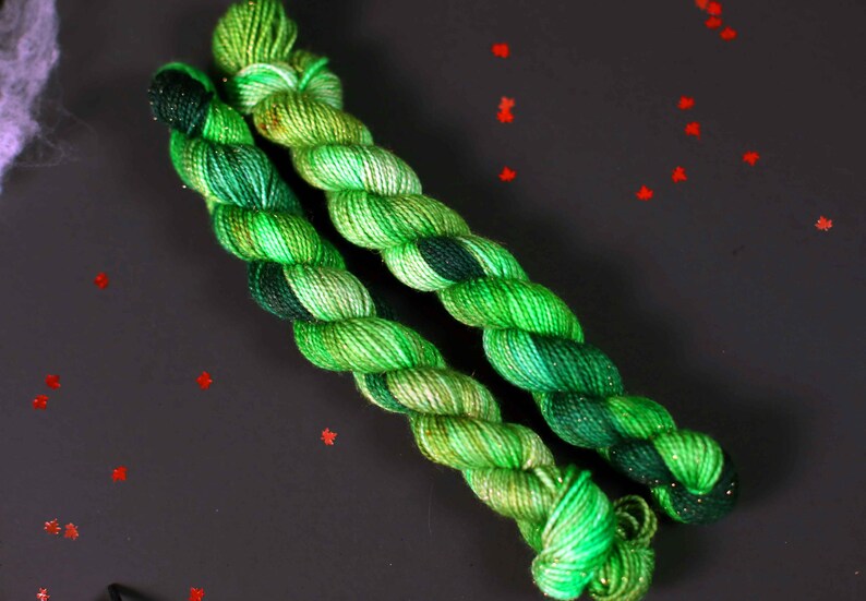 May include: Two skeins of green yarn with a speckled pattern. The yarn is twisted together and has a dark green and light green colour variation.