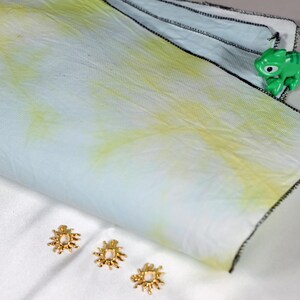 May include: A folded white and light blue fabric with yellow accents and black stitching. A small green toy lizard sits on the fabric. Three gold-coloured decorative flower-shaped objects are also present.