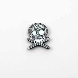 May include: A silver-colored enamel pin in the shape of a skull with a yarn ball design and crossed needles. The skull has white eye sockets and a detailed design. The needles are crossed below the skull.