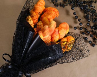 May include: A bouquet of orange and yellow yarn wrapped in black netting and paper, tied with a black ribbon. Dark berries and foliage are arranged on a brown surface, complementing the yarn's colors. The yarn is a vibrant mix of orange and yellow.