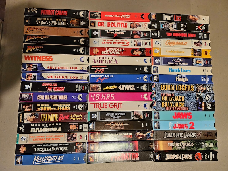 May include: A collection of VHS tapes featuring various movie titles, including "Patriot Games", "Six Days Seven Nights", "Indiana Jones", "Raiders", "Witness", "Air Force One", "The Fugitive", "Clear and Present Danger", "The Sum of All Fears", "John Wayne and Duke Westerns", "Ransom", "Lethal Weapon", "Tequila Sunrise", "Hellfighters", "Dr. Dolittle", "Coming to America", "Beverly Hills Cop", "48 Hrs.", "True Grit", "Conan", "Predator", "True Lies", "The Running Man", "Caddyshack", "Fletch Lives", "Fletch", "Born Losers", "The Trial of Billy Jack", "Jaws", "Jaws 2", "Jurassic Park", "The Lost World", and "Jurassic Park".