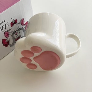May include: White ceramic mug with a pink paw print design. The mug is shaped like a cat's paw and features a cartoon image of Marie from The Aristocats on the side. The text "Disney Marie" is printed on the box.
