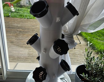 Op de afbeelding: Witte plastic hydroponische toren met zes zwarte plantenpotten, een witte emmer met blauwe rand en een zwart snoer. De toren staat op een wit oppervlak.