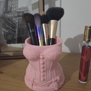 Peut inclure: Un porte-pinceaux rose en forme de corset contenant plusieurs pinceaux de maquillage. Le support présente des détails décoratifs et des nœuds. Un flacon de parfum est à droite et une photo en noir et blanc encadrée est en arrière-plan.