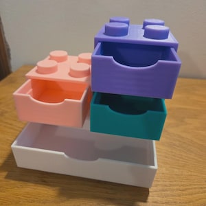 Peut inclure: Tiroirs de rangement empilables et colorés en forme de blocs de construction. Les tiroirs sont rose pastel, violets, turquoise et blancs. Chaque tiroir est coulissant et en plastique. Les tiroirs sont empilés les uns sur les autres.