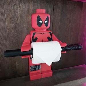 Può includere: Una figura in plastica rossa a forma di personaggio a blocchi con il design della maschera di Deadpool, caratterizzata da dettagli neri sugli occhi e sul busto. La figura regge una lunga asta cilindrica nera che sostiene un rotolo di carta igienica.