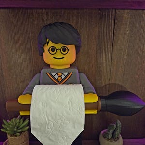 Puede incluir: Un original portarrollos de papel higiénico con forma de figura estilo Lego con una escoba. La figura tiene pelo negro, gafas y un atuendo gris y amarillo. La escoba sostiene un rollo de papel higiénico blanco. Pequeñas plantas en macetas a ambos lados.