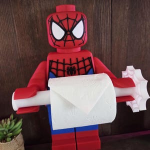 Op de afbeelding: Een rood en blauw plastic figuur ontworpen als een blokachtig personage met een Spider-Man-masker, die een witte horizontale stang vasthoudt die een rol wit toiletpapier ondersteunt. Het figuur heeft zwarte webpatronen op het hoofd en de borst.