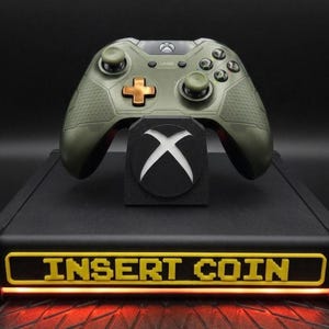 Könnte beinhalten: Ein olivgrüner Xbox-Controller mit einem goldfarbenen Steuerkreuz liegt auf einem schwarzen Ständer. Der Ständer hat ein weißes Xbox-Logo und steht auf einer schwarzen Basis mit den gelben Worten "INSERT COIN". Die Basis hat ein rotes Unterlicht.