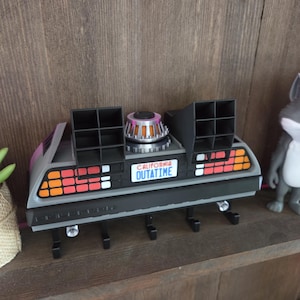 Puede incluir: Un llavero DeLorean gris y negro con luces naranjas y blancas, y la matrícula dice "California OUTATIME". El llavero tiene cuatro ganchos negros y un centro rosa y plateado.