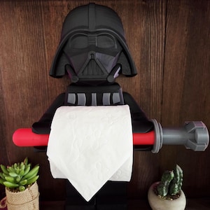 Könnte beinhalten: Ein schwarzer Darth Vader Toilettenpapierhalter. Der Halter hat eine rote Lichtschwert-Stange, um die Toilettenpapierrolle zu halten. Das Design umfasst einen schwarzen Helm und Körper, mit einer Rolle weißem Toilettenpapier.